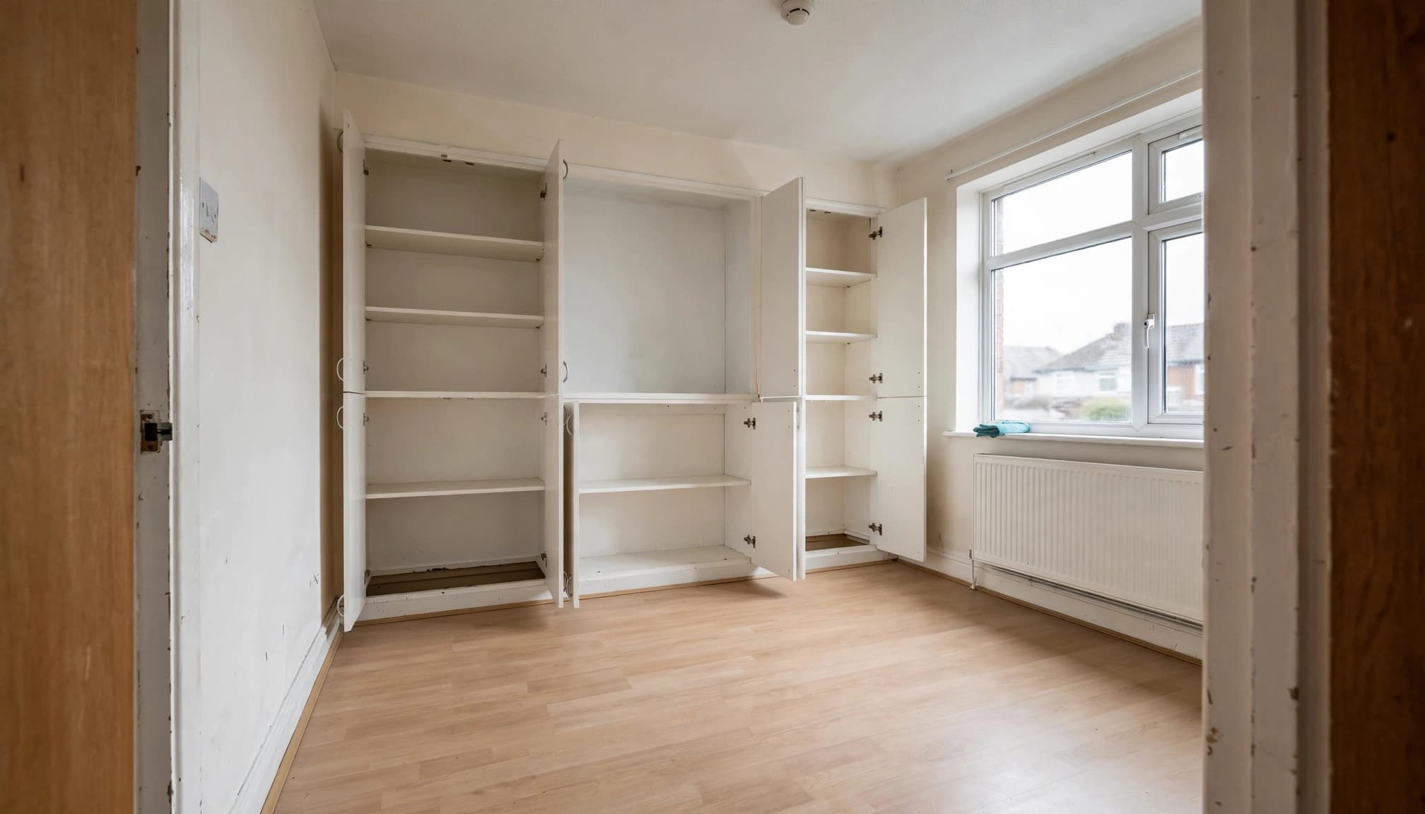 An empty UK room with laminate flooring, open white built-in cupboards, a radiator, and a window.